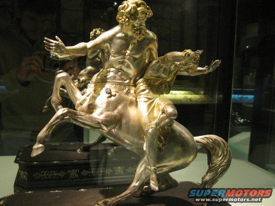 Silver sculpture of a centaur riding a horse, with ornate base. Reflection of photographer visible