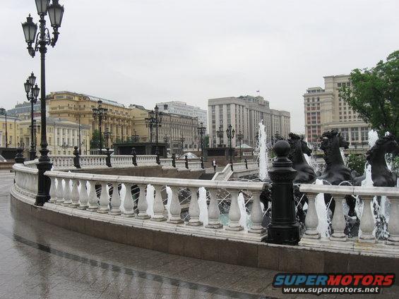 Fountain with horse sculptures and ornate railing in front of buildings under a cloudy sky