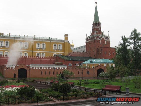 Kremlin buildings with a red tower and yellow facade, steam rising from a gate, and a park with benches