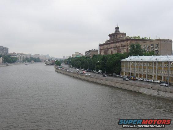 Cityscape with a wide river, a tree-lined embankment, and buildings under a cloudy sky. Cars are parked along the road