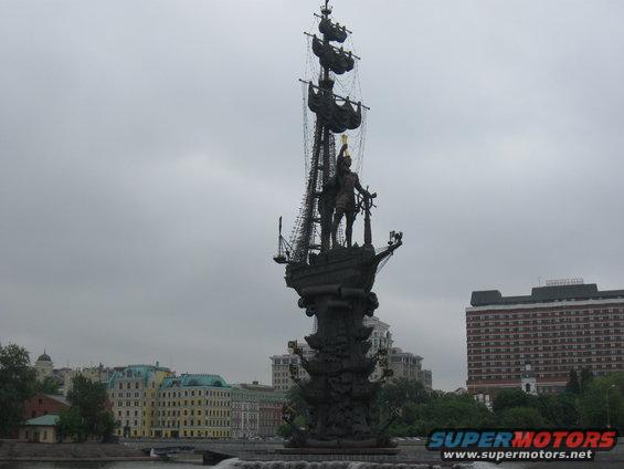 Monument of Peter the Great with ship masts, against a cloudy sky, with buildings and trees in the background