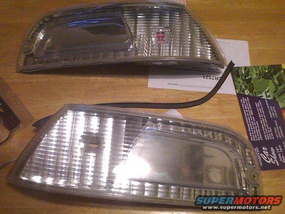 Two clear plastic car headlights with ribbed lenses, one above the other, on a wooden surface