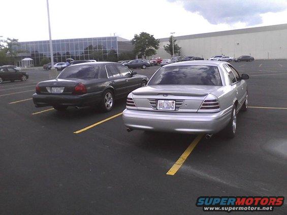 Two Ford Crown Victorias parked in a lot. One is silver, the other is dark green. The silver one has a license plate that reads NBL 3645