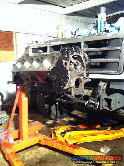 Engine block on an orange engine hoist in front of a vehicle's silver grille and front end