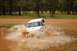 1996 Ford Bronco - A white 1996 Ford Bronco kicks up a large spray as it drives through a muddy puddle in a wooded area.