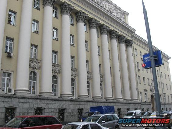 Building with large columns, cars parked on street, blue tarp covering object