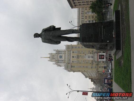 Statue of a man in a suit on a pedestal, with a tall building and city street in the background