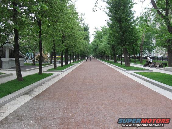 Long park path lined with trees, people walking in distance. Supermotors logo in corner