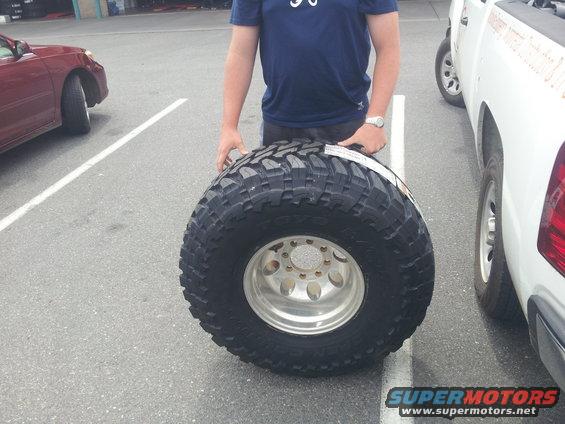 Person holding a large off-road tire and wheel. White pickup truck and red car visible in background
