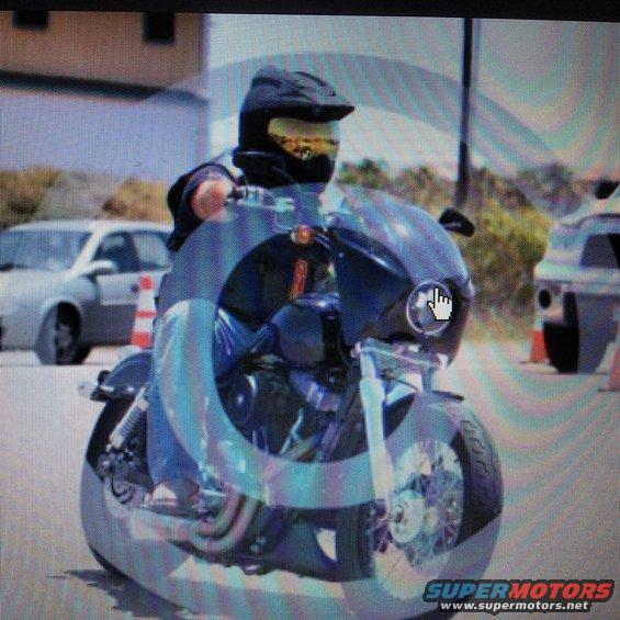 Motorcyclist wearing a helmet and goggles rides a dark motorcycle with a fairing. Cars are visible in the background