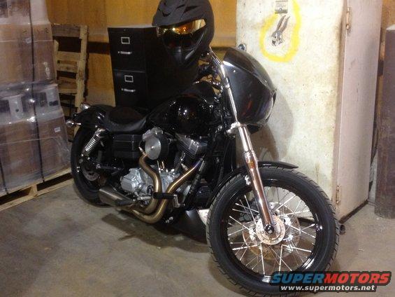 Black motorcycle with chrome spokes and exhaust, parked indoors next to a filing cabinet and pallets
