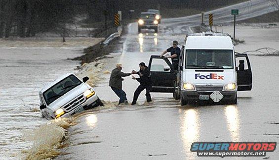 White Ford Ranger tilted in floodwater, driver's side door open, with people assisting