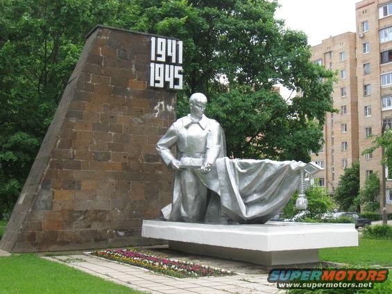 Monument with dates 1941-1945 and a silver statue of a man in front of a brick structure and apartment buildings