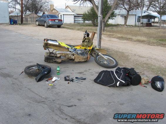 Dirt bike on its side with tools, a helmet, and bags laid out on the pavement