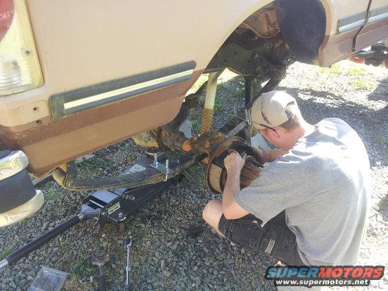 Guy working on the rear axle and suspension of a tan truck. It looks like a Ford Bronco