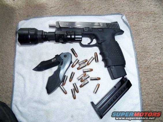 Smith & Wesson M&P pistol with mounted flashlight, a folding knife, loose bullets, and a detached magazine on a white cloth