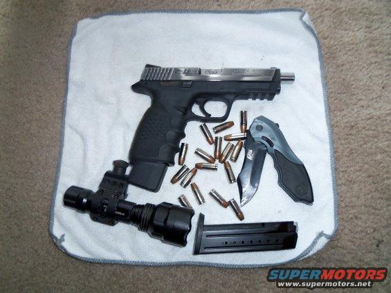Pistol with extended magazine, loose rounds, a folding knife, and a tactical flashlight laid out on a white cloth