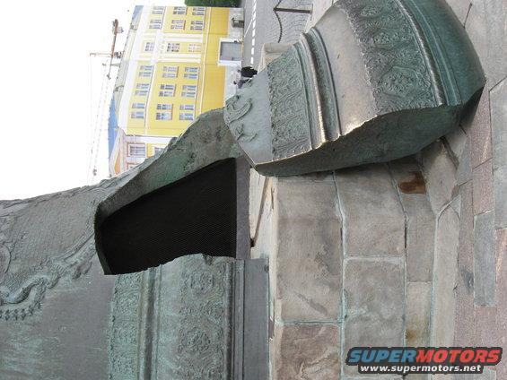 Close-up of a large, ornate bronze sculpture with a yellow building in the background. Supermotors.net logo visible