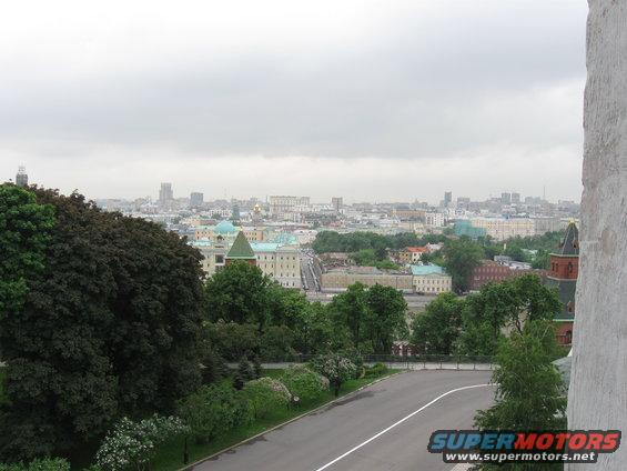 Cityscape view with buildings, trees, and a road in the foreground. Watermark reads SUPERMOTORS