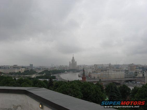 City skyline with a river, trees, and a prominent tall building under a cloudy sky. Supermotors.net logo in corner