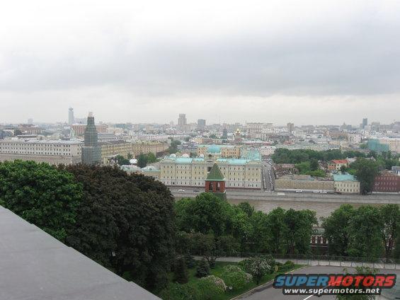 Cityscape with numerous buildings, a wide river, and trees under a cloudy sky. A watermark reads SUPERMOTORS