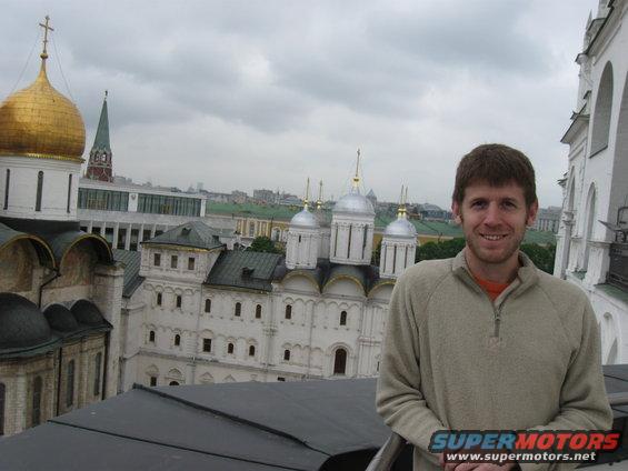 Man in tan quarter-zip sweater smiles in front of golden-domed Kremlin cathedrals under a cloudy sky