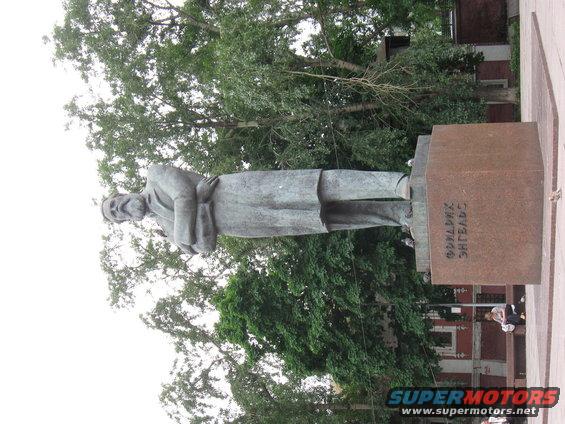Stone statue of a man with arms crossed, on a pedestal with Cyrillic text, outdoors with trees and buildings