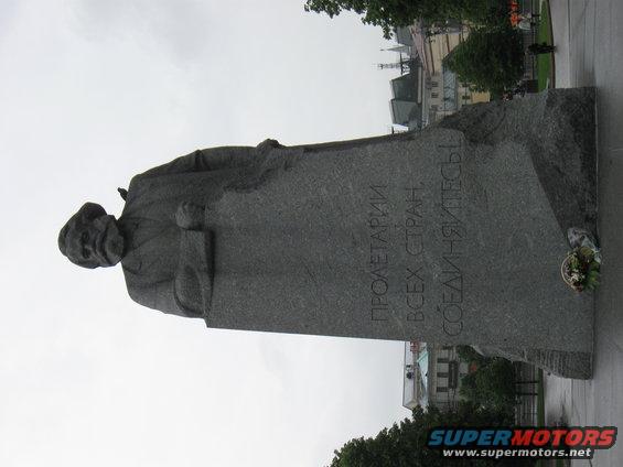 Large stone monument with text in Russian, buildings and trees in background