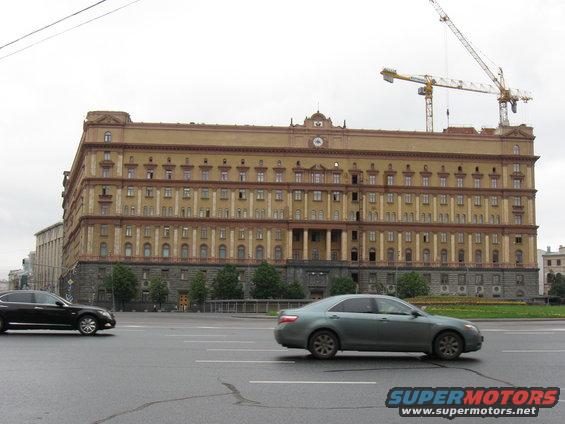 Gray sedan and black sedan on wet road in front of large tan building with cranes overhead