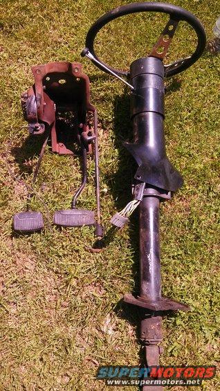Steering column and pedals on grass. Steering wheel has holes. Wiring harness visible