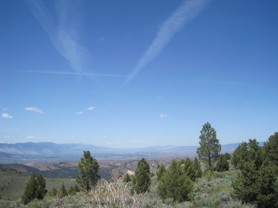 View of a valley and mountains under a blue sky with contrails, foreground has trees and brush