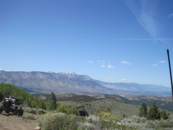 Two dark-colored off-road vehicles on a dirt path, one in the foreground on the left, the other further back on the right, with a vast mountain range under a clear blue sky