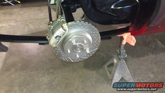 Rear brake rotor with drilled holes and silver caliper, part of a vehicle suspension system on jack stands