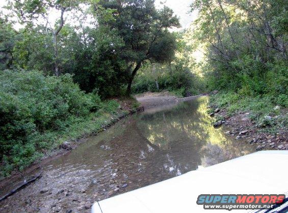 White vehicle's hood and windshield driving through a shallow, rocky stream in a wooded area