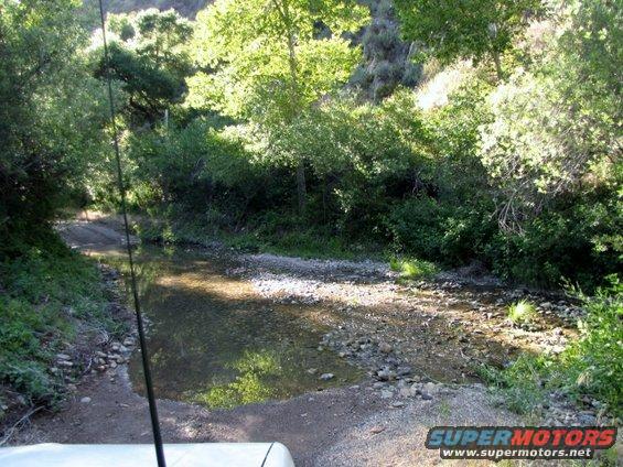View from inside a vehicle crossing a shallow, rocky stream in a wooded area