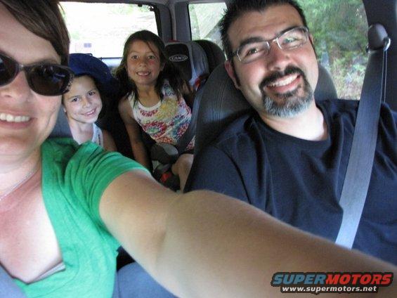 Family taking a selfie inside a vehicle. A woman and two children are in the front and middle rows, with a man in the driver's seat