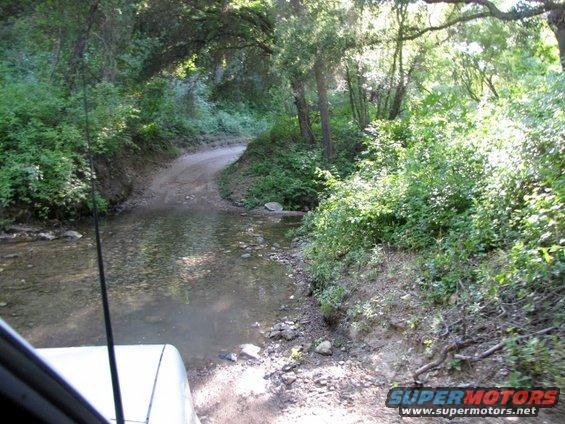 View from inside a vehicle driving on a dirt road that crosses a shallow stream with trees and brush on either side