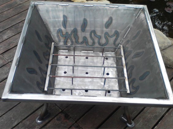 Square metal fire pit with grate inside, sitting on wooden deck. Flame shapes cut into the inner walls