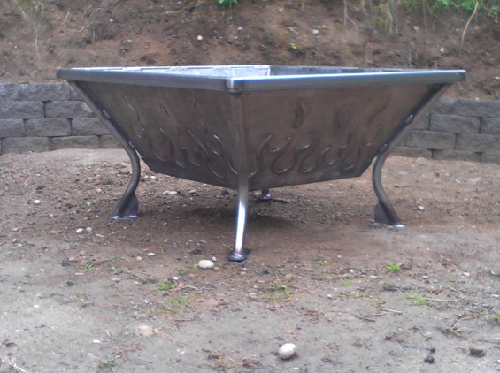 metal fire pit with flame cutouts on legs, set on dirt ground near a stone wall