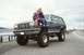 1979 Ford Bronco - A red Ford Bronco is loaded onto a trailer, with a black truck hitched to it, in a wooded area.