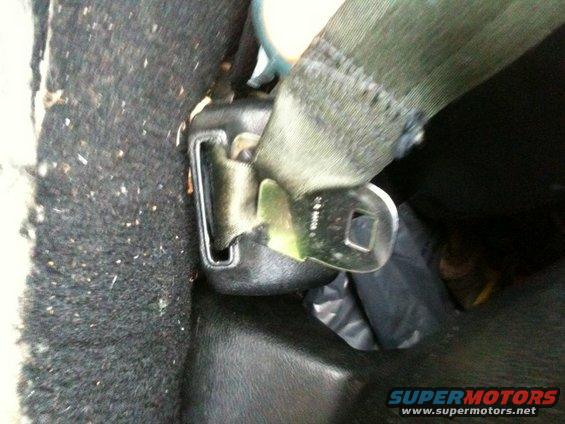 Seatbelt buckle with green strap and metal clasp, attached to dark carpet and black plastic trim