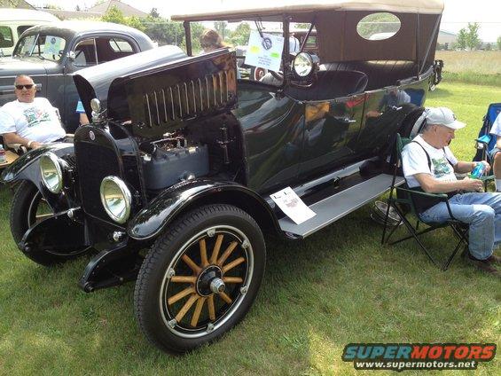 Black touring car with hood open, two men sitting in chairs nearby. Another car is parked behind it
