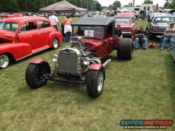 Red hot rod with exposed engine and large rear tires parked on grass