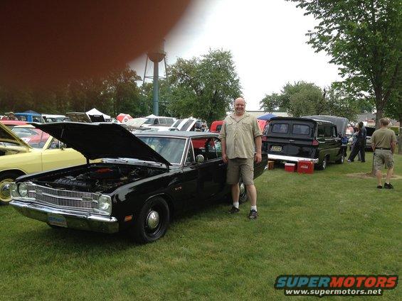 Guy standing next to a black classic car with its hood up at an outdoor car show. Other cars and a water tower are in the background
