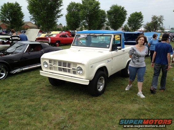 White 1973 Ford Bronco with blue hardtop parked on grass, other cars visible in background