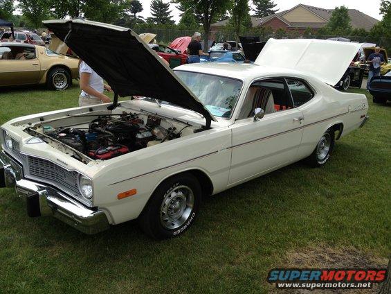 White coupe with hood open, showing engine. Chrome bumper, red stripe on side. Other cars visible in background