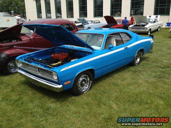 Blue 1973 Plymouth Duster coupe with white side stripes, hood open showing engine. Other classic cars visible