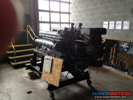 Large V12 engine on a rolling stand in a museum or workshop. Display board to the right has text about automakers