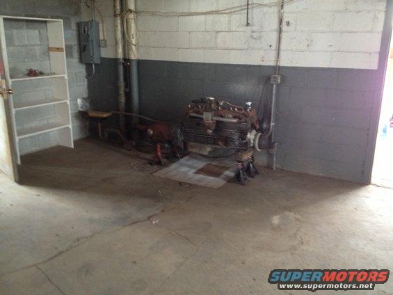 Engine and transmission assembly on jack stands in a garage. Shelving unit on left