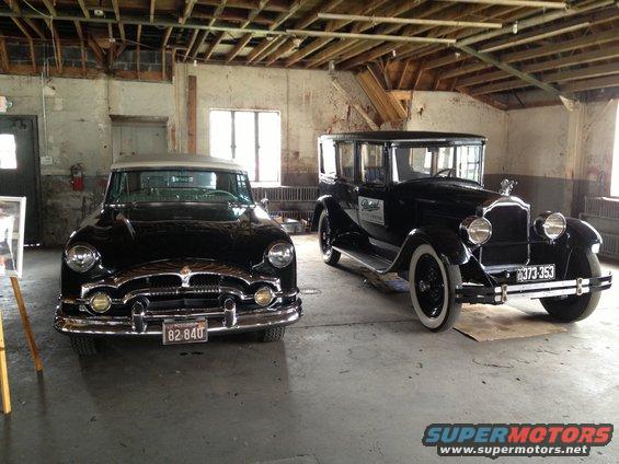 Two old black cars parked inside a garage. The one on the left has a chrome grille and the one on the right has a tall grille